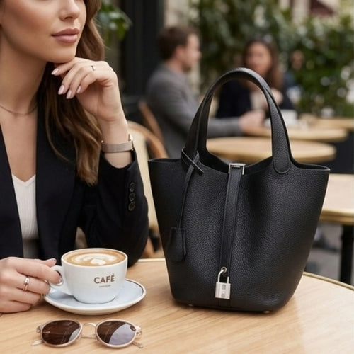 Une femme élégante est assise à une table de café, tenant une tasse. Sur la table se trouve son sac à main noir en cuir, son smartphone et ses lunettes de soleil.