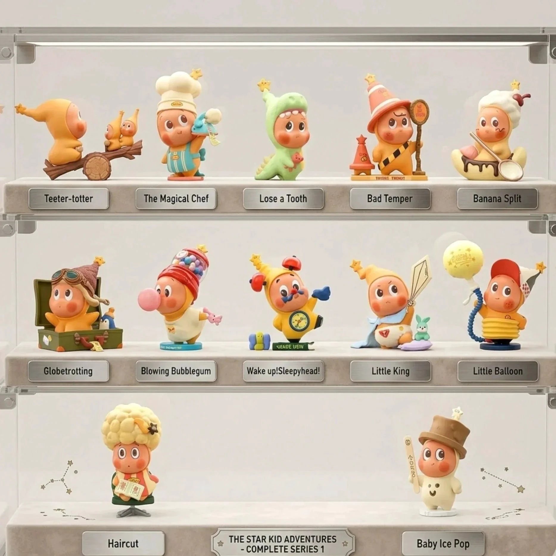 Une vitrine de collection transparente en acrylique à trois étagères contenant la collection 'The Star Kid Adventures - Complete Series 1'. Chaque étagère présente des figurines en vinyle distinctes avec des étiquettes individuelles en métal comme 'Teeter-totter', 'Globetrotting', et 'Little King'. L'étagère du milieu contient la figurine 'Little Balloon'. Les étagères sont éclairées par des lumières LED douces et intégrées. L'arrière-plan de la vitrine présente un motif de constellation subtil.